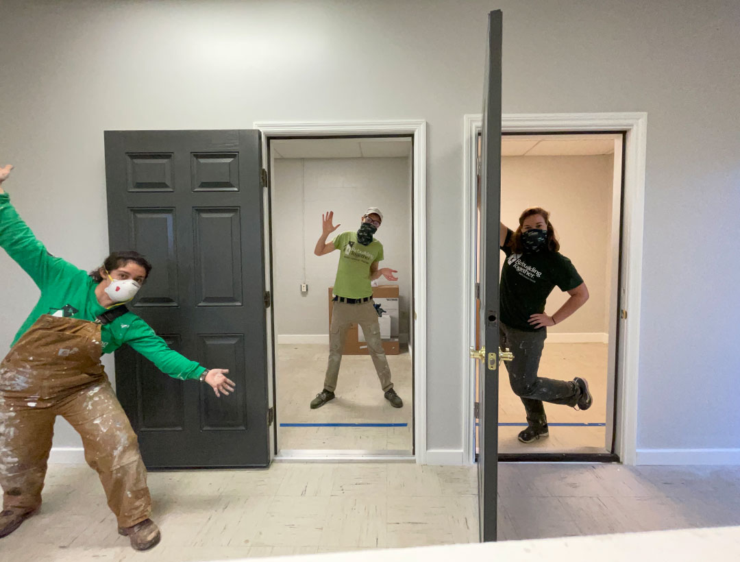 R.D., masked, standing and posing in a door frame between the two current AmeriCorps members on either side of him. One member is posing with arms outstretched to sides and the other is leaning against another door frame. They are all inside an empty office space.